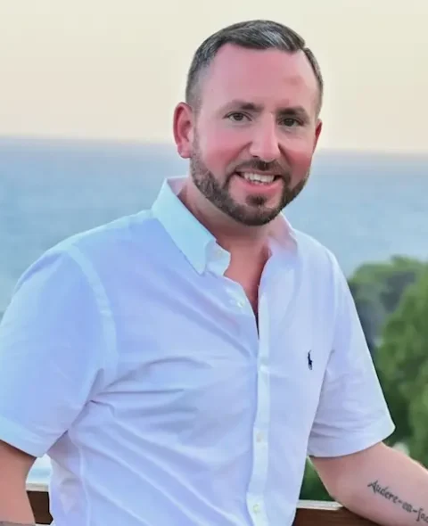 Chris Meaney stands outdoors, smiling in a white button-up shirt, with the ocean and trees forming a serene backdrop.