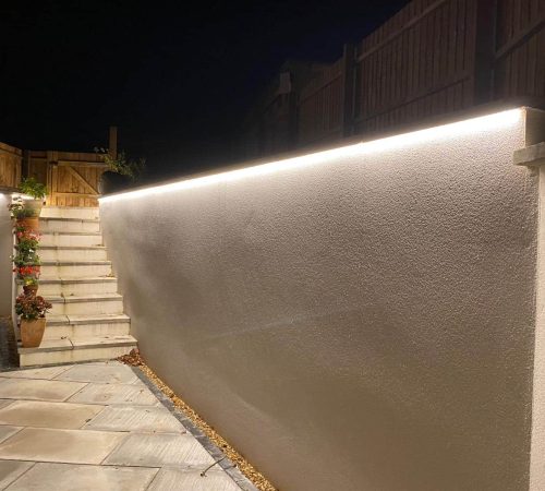 A nighttime outdoor scene at Tinwick Crescent reveals a stone patio with a white wall illuminated by a strip of bright lights along the top. Adjacent are steps leading up, bordered by potted plants, and a wooden fence above.