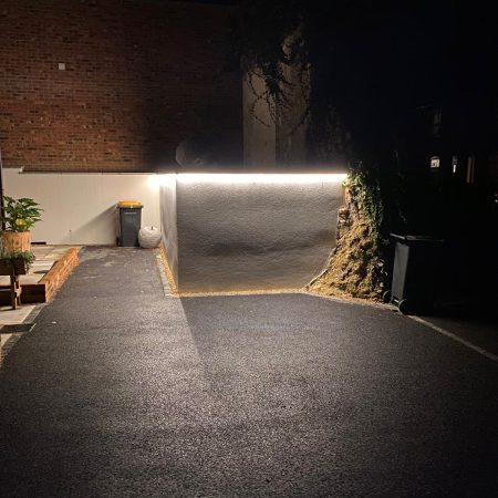 A dimly lit alley at night reminiscent of The Barn, featuring a brick wall and a white wall illuminated by a bright strip light. A trash can and potted plants are visible, while the asphalt ground lies partially in shadow, offering stark contrast to the illuminated area.